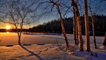 A serene winter sunset casting shadows on a frozen lake surrounded by snow and trees.