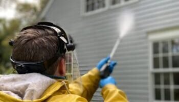 Deck Cleaning Person cleaning house siding with a pressure washer, wearing protective gear in a yellow raincoat.