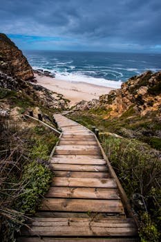 Wooden stairs leading down to a scenic beach surrounded by cliffs under a dramatic sky.